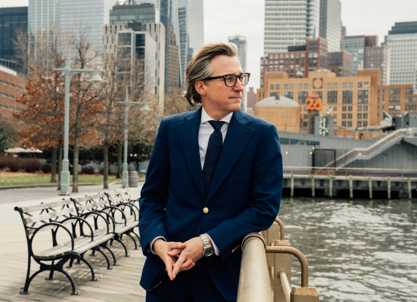 Doctor Craig Baldenhofer MD in navy suit sitting on bench by waterfront with city skyline background - in New York, NY