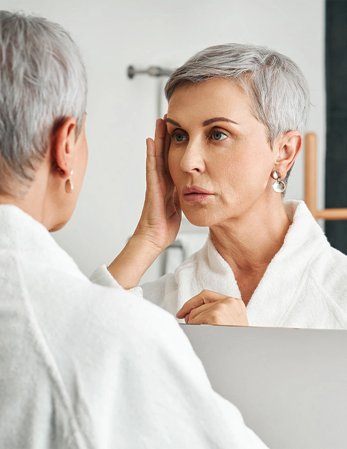 Mature woman with short gray hair examining her face in bathroom mirror while wearing white bathrobe - Facelift in New York, NY