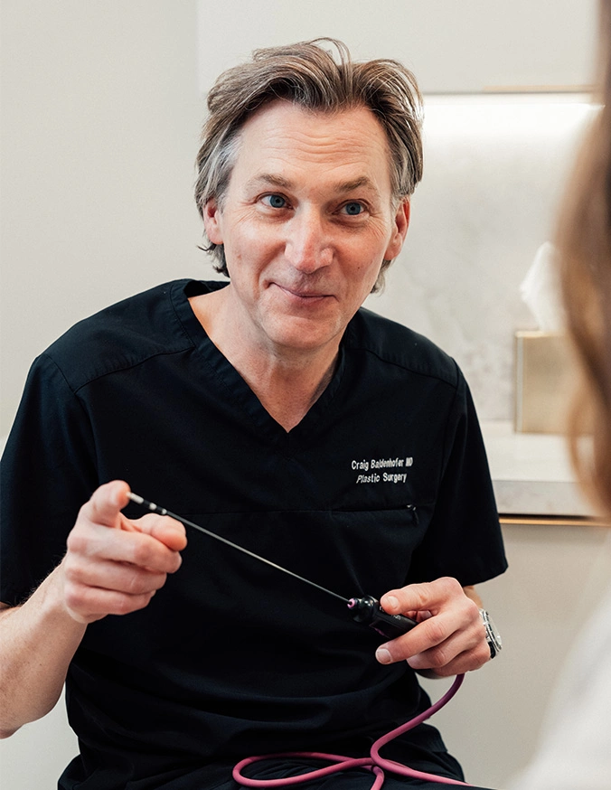 Doctor Craig Baldenhofer MD in black scrubs holding surgical instrument while explaining procedure to patient - Fat transfer breast augmentation in New York, NY