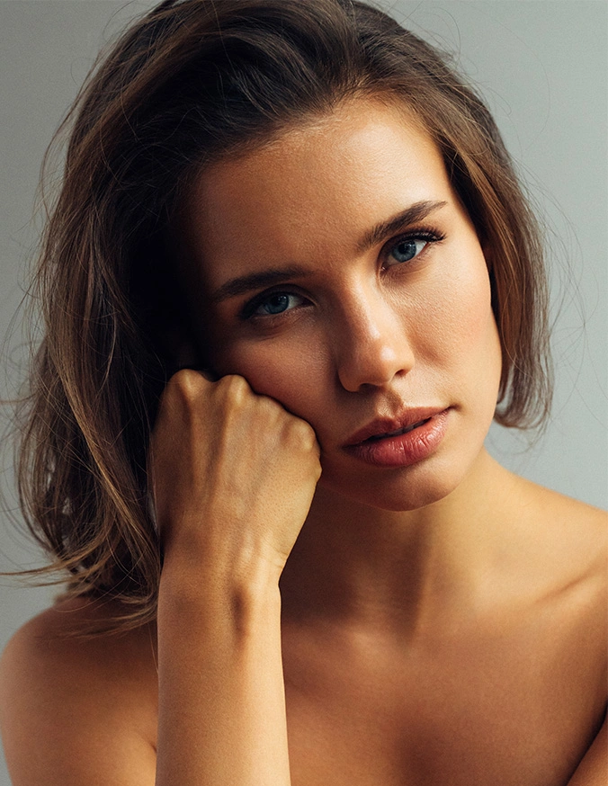 Thoughtful young woman with shoulder-length brown hair resting face on hand against gray background - Fat transfer to face in New York, NY