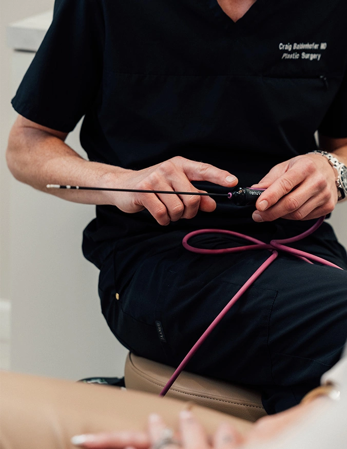 Dr Craig Baldenhofer MD holding medical device and showing it to patient laying down. - Mommy makeover in New York, NY