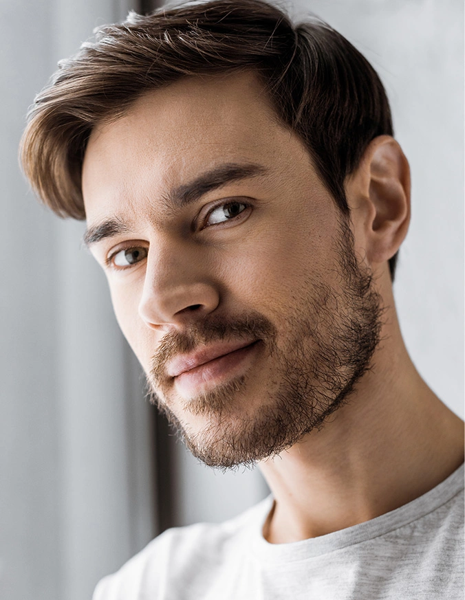 Handsome young man with brown hair and beard looking at camera with confident smile against light background - Otoplasty in New York, NY