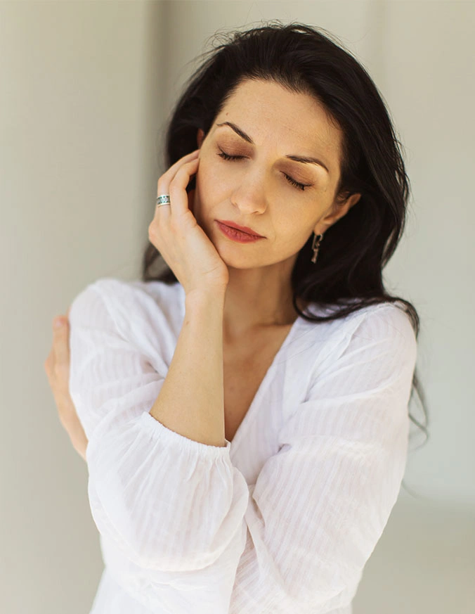 Woman in white blouse with hand on face showing thoughtful, serene expression with eyes closed - Upper eyelid surgery in New York, NY