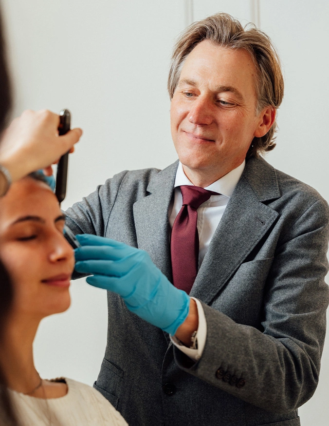 Doctor Craig Baldenhofer MD smiling as he applies product to the patients face and wearing blue gloves. - Alloclae in New York, NY