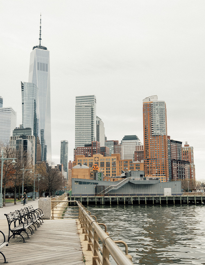 Manhattan skyline view from waterfront walkway with benches and One World Trade Center tower - in New York, NY