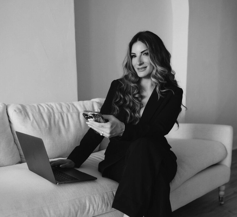 Professional businesswoman in black suit sitting on white couch with laptop and coffee during remote work - in New York, NY